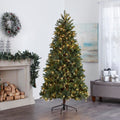 Hudson Spruce Christmas Tree with Warm White Lights, in Living Room In Front of Fireplace. Fireplace Decorated with Garland and Wreath Above on Wall, Giftboxes to Right of the Tree on Ottoman.