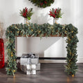 Mini Pinecone Garland Decorating Front View of Table. Silver Boxed Presents Underneath Table. Potted Plant Decor on Top of Table. Partially Visible Wreath on Wall Above. White Wall Background.