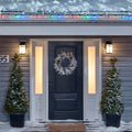 Noma C6 Multi-Color String Lights on a roof edge above home entrance. Wreath on door and two potted trees on porch