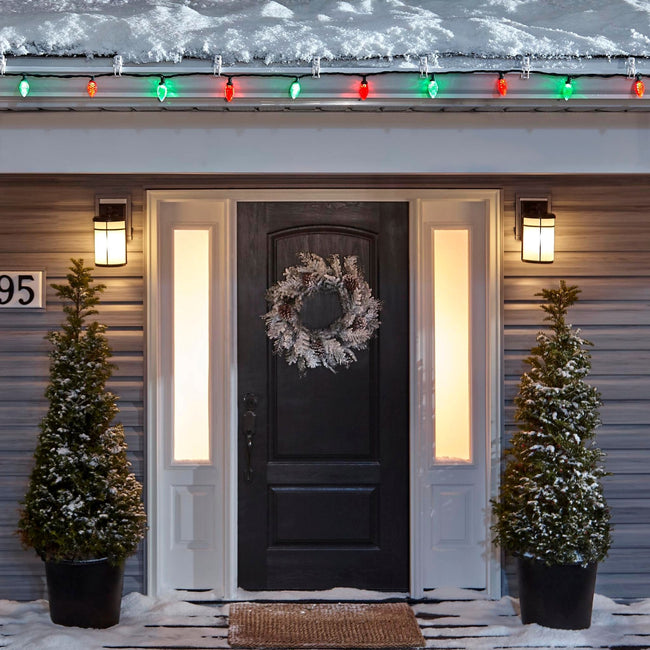 Noma C9 Red & Green String Lights, on a roof edge above home entrance. Wreath on door and two potted trees on porch